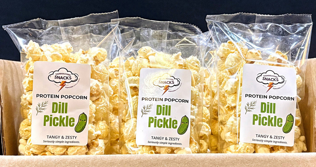 Three bags of Dill Pickle Protein Popcorn in a cardboard box with a dark background