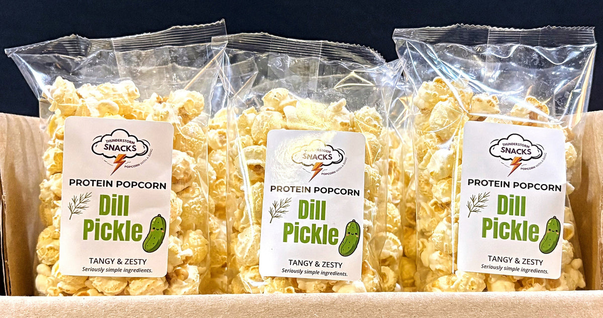 Three bags of Dill Pickle Protein Popcorn in a cardboard box with a dark background